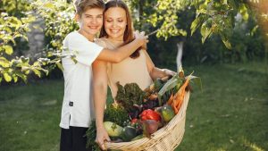 Mother and son smiling outdoors with a basket full of fresh vegetables, embracing in a sunny garden.